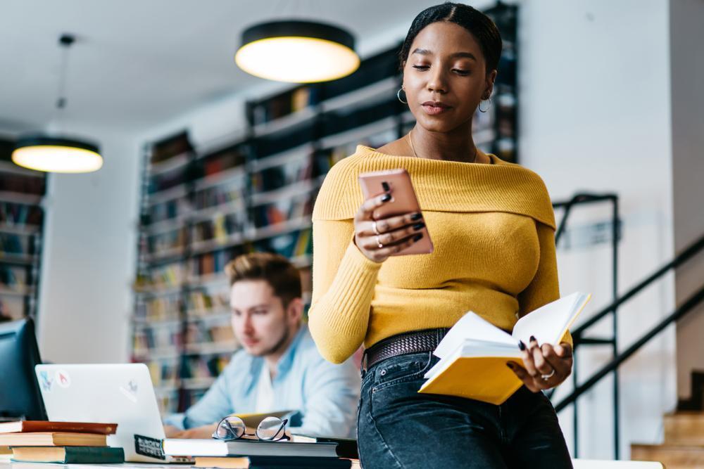 beautiful black woman looking at smartphone held in right hand with open book in left hand  resting on pelvic area leaning on table in library white male sitting at table in background reading a laptop screen reading glasses resting on table